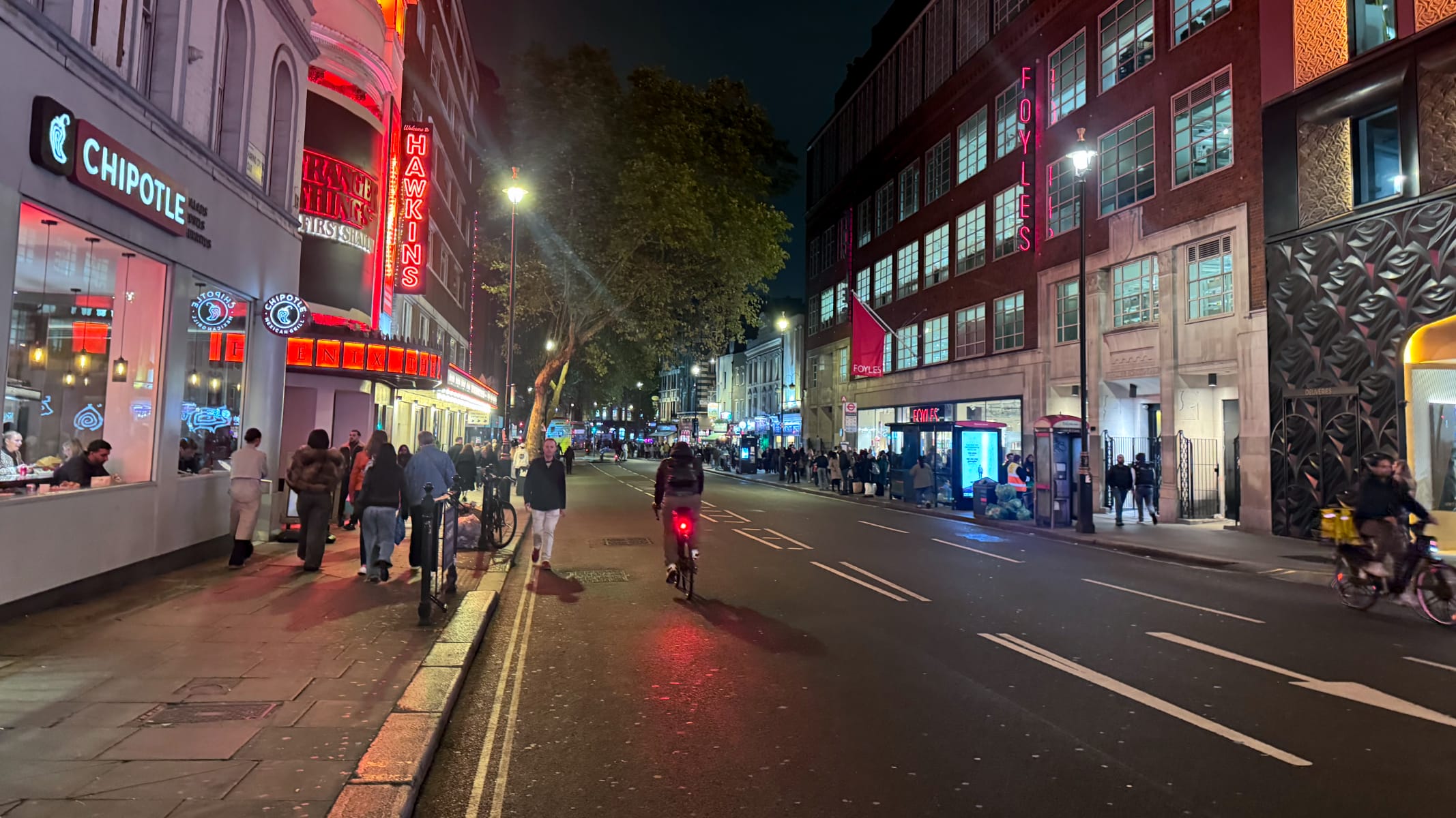 London's Charing Cross Road at night with packed sidewalks but a nearly empty road, with only a few bicycles on it.