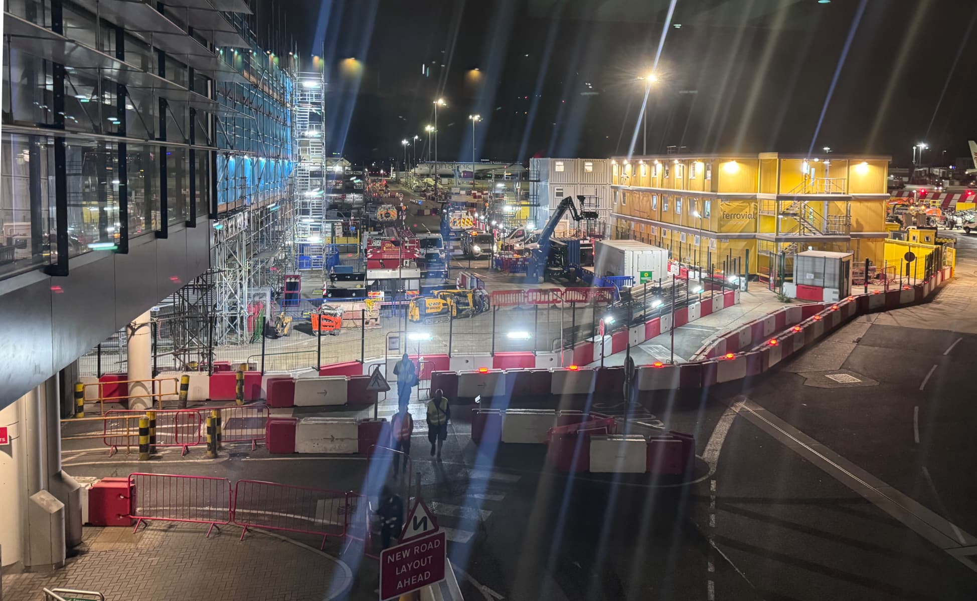 A night view of a construction rallying area at Heathrow Terminal 2, with stacked prefab units, scaffolding, and orange barriers reorienting traffic. The lights are smudged and there is window reflection glare.