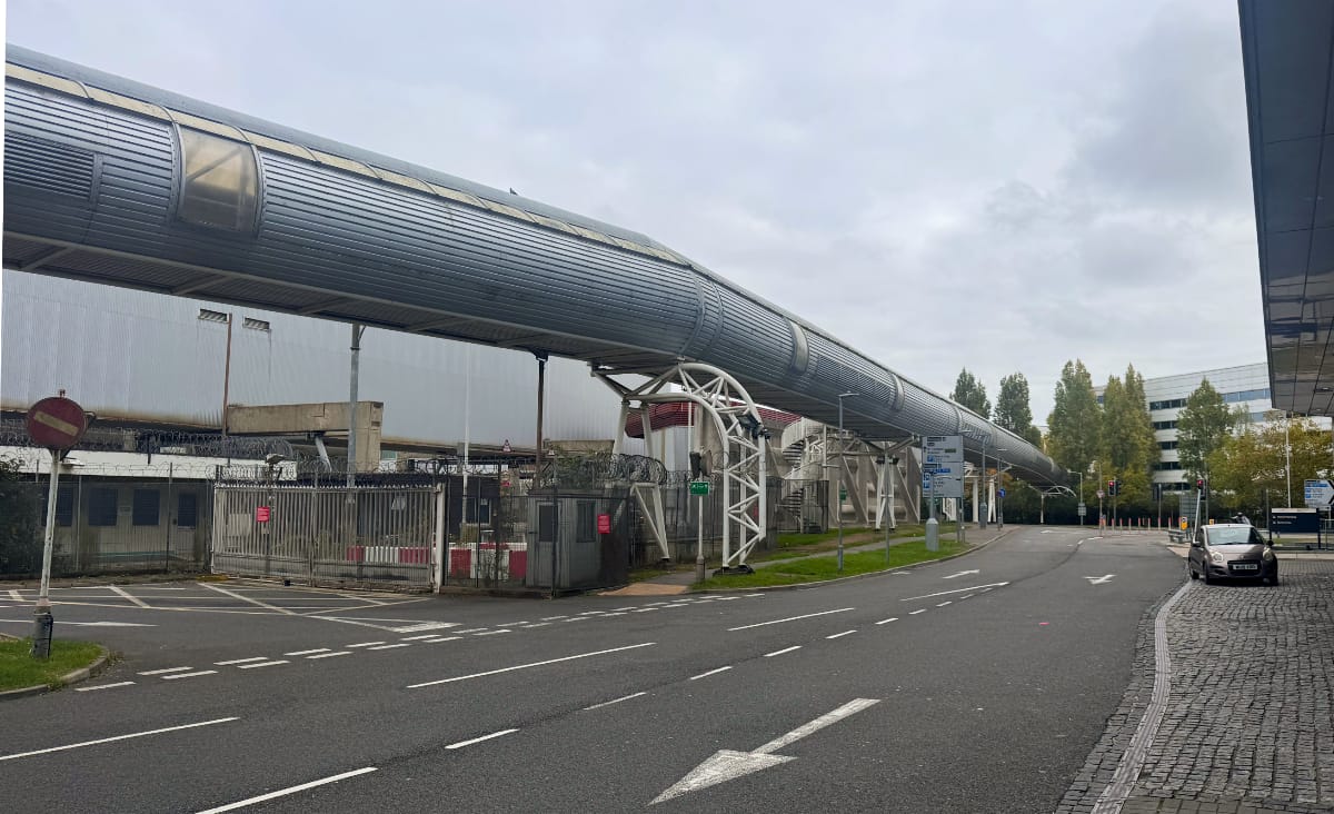 An exterior view of a roadway and buildings, with a scruffy raised walkway tube above