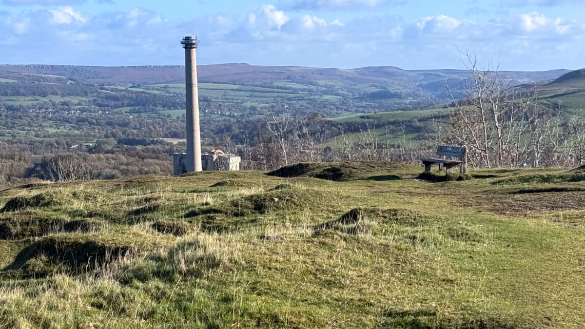 A rugged field with a smokestack in the distance and a lone bench