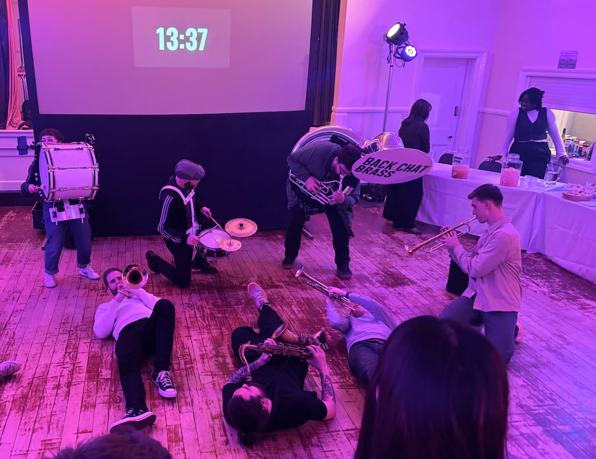 The Back Chat Brass Band playing sprawled on the floor under purple lights, with a countdown timer on a screen behind reads 13:37