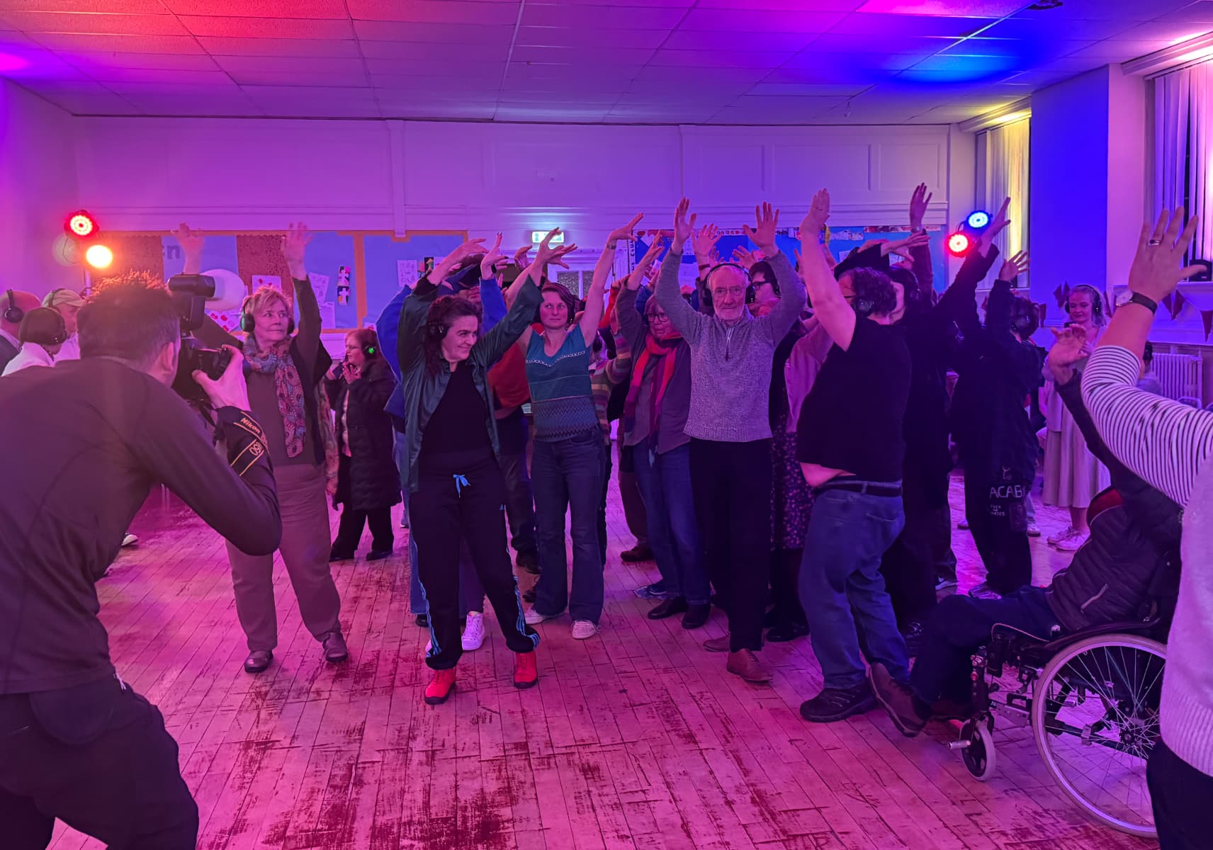 A large and varied group of participants doing dance-like moves in a purple-lit room