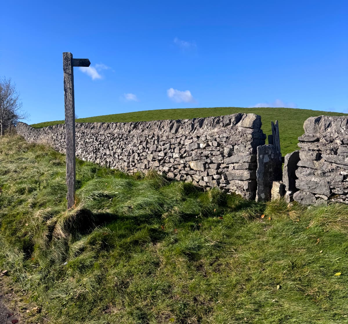 A wooden signpost pointing to a gap in a stone fence on a hillside