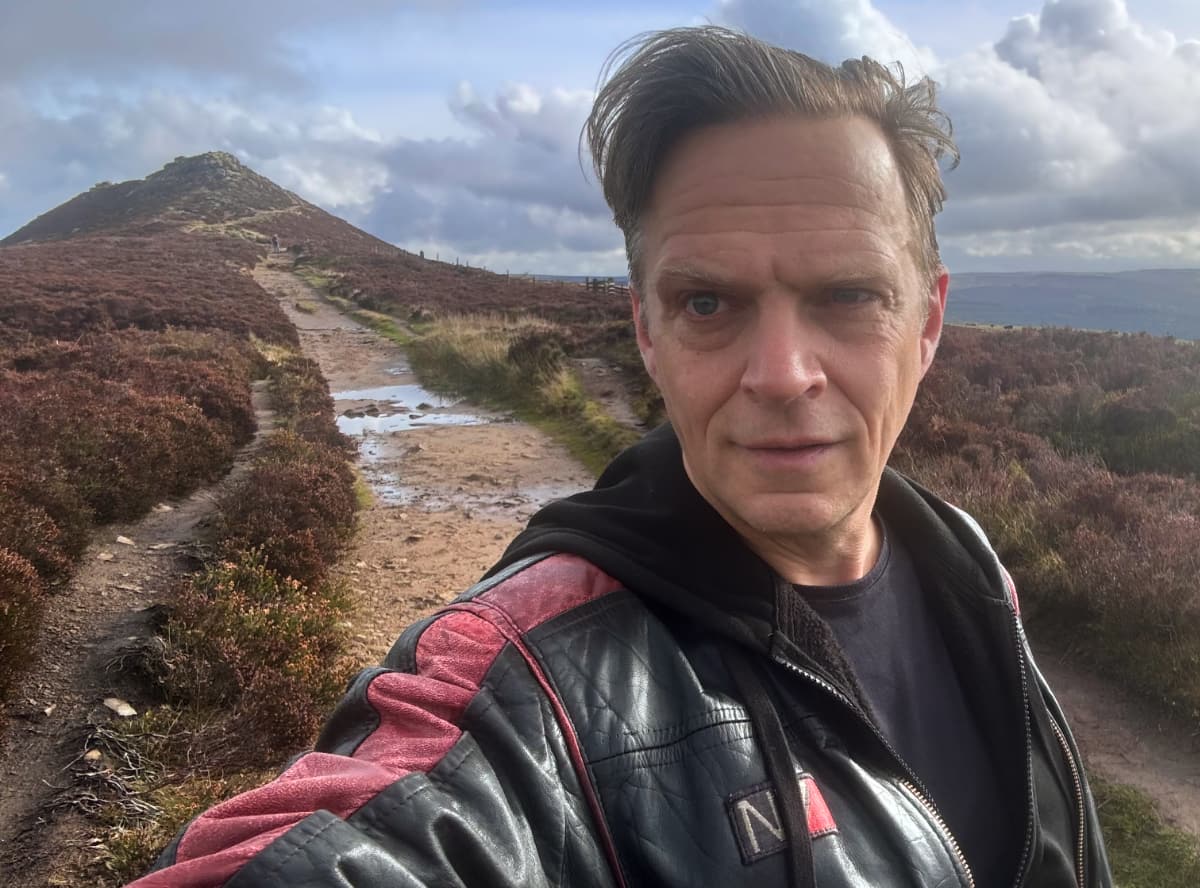 A selfie of Andrew Burke in a leather N7 jacket, with the top of Win Hill behind