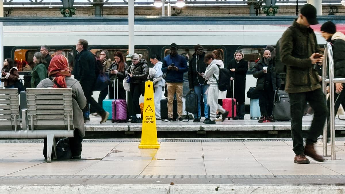 A widescreen zoom shot of people waiting on a train platform at Manchester Piccadilly, mostly looking at their phones