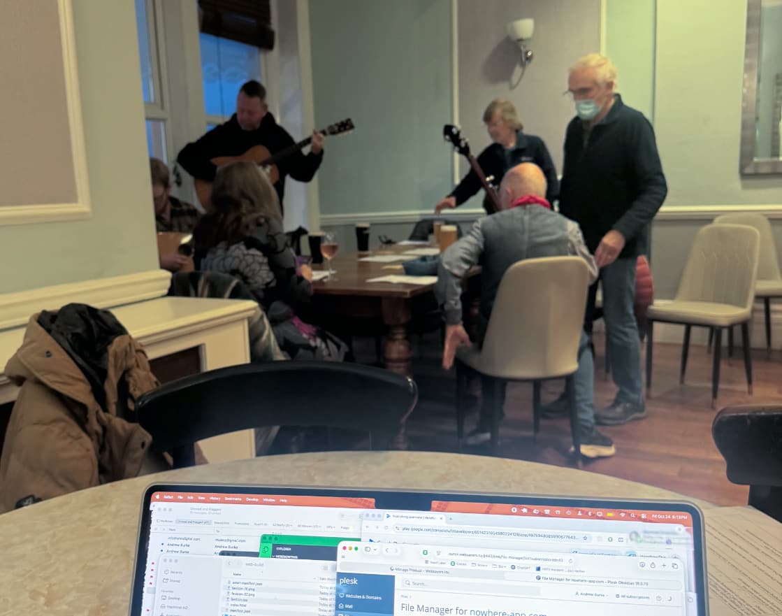 A group of musicians gather around a table while in the foreground is the top of a laptop screen with many windows, including email, terminals, and a browser pointing to a Plesk interface